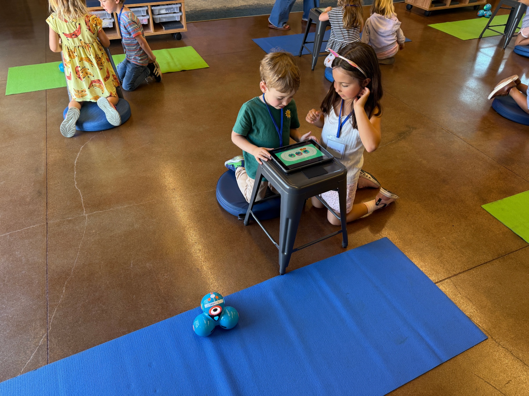 Kindergarten students working together on a tablet to program a small blue robot on a blue mat.